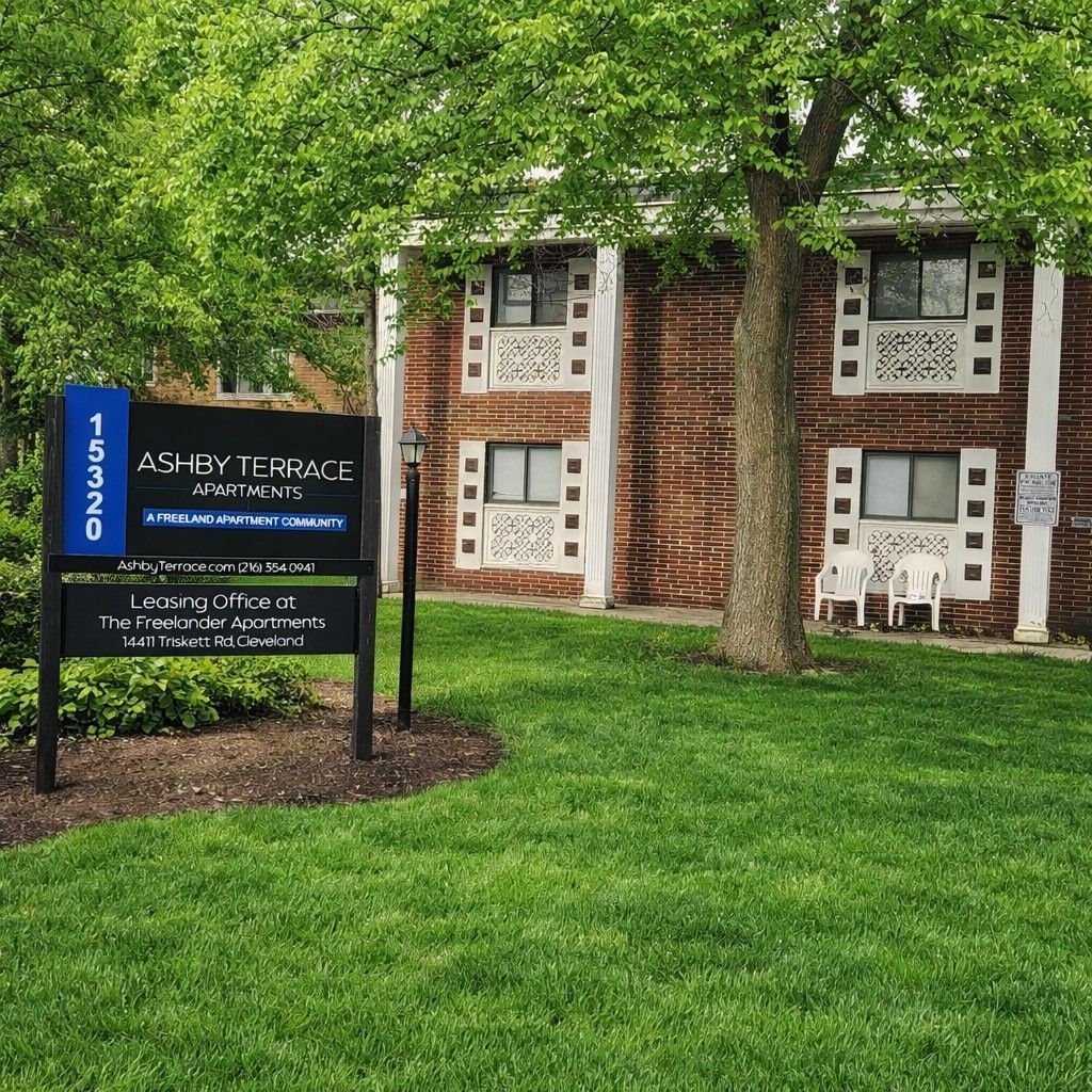 A sign for Ashley Terrace Apartments stands on a grassy lawn in front of a two-story brick building with white trim.