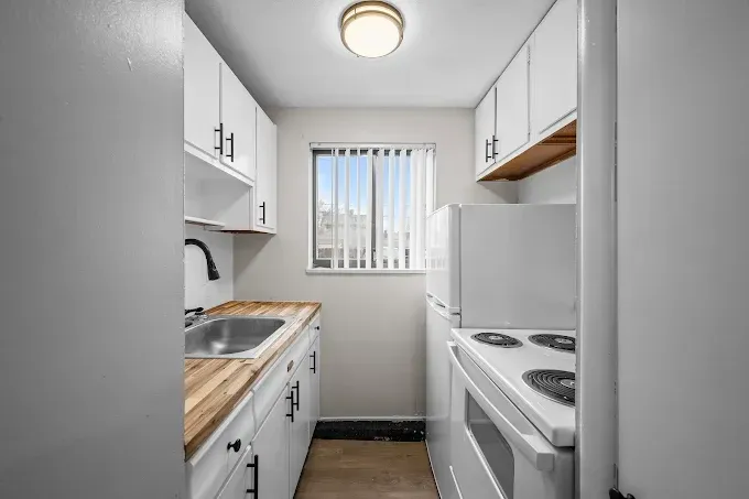 A compact, galley-style kitchen with white cabinets, a light wood countertop, a stainless sink, and white appliances.