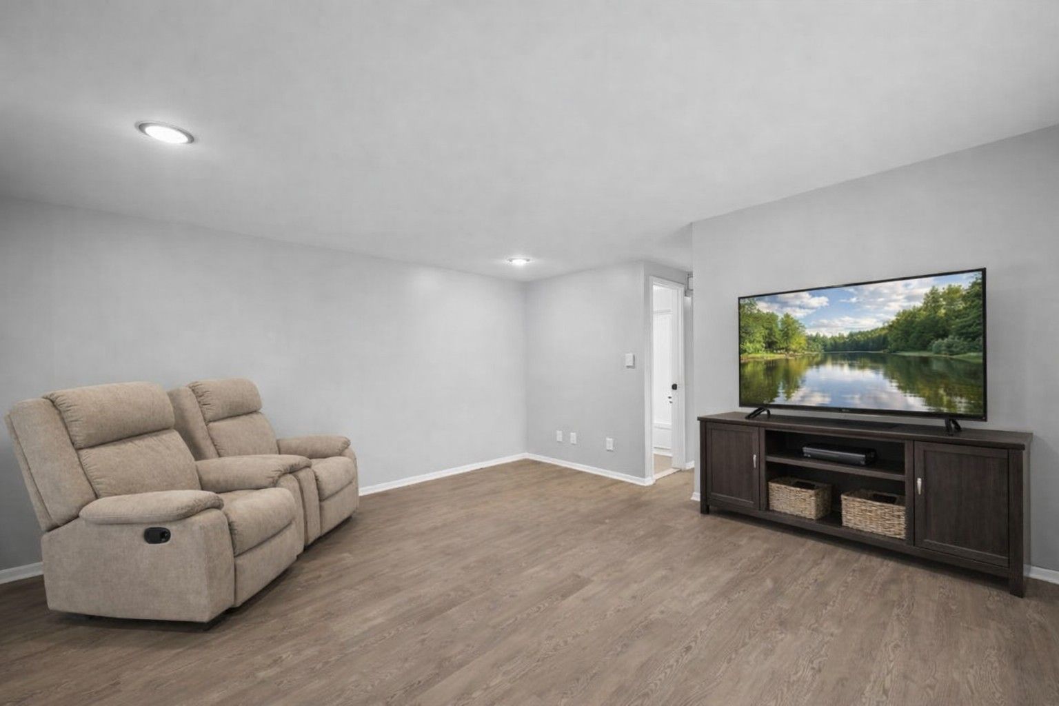 A beige two-seat recliner faces a television on a dark wood media console in a room with light grey walls and wood floors.