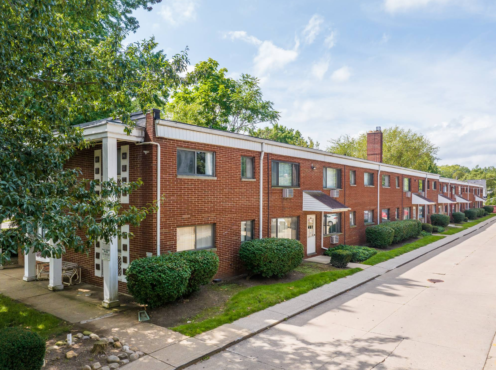 A long, two-story red brick apartment building with white columns and trimmed green bushes along a paved road.