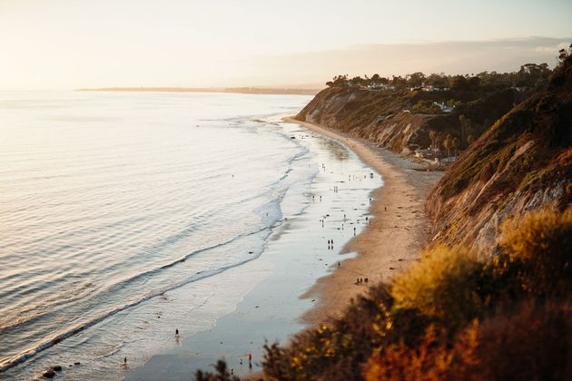 Sunlit beach with gentle waves, rocky cliffside, and a coastal path at sunset