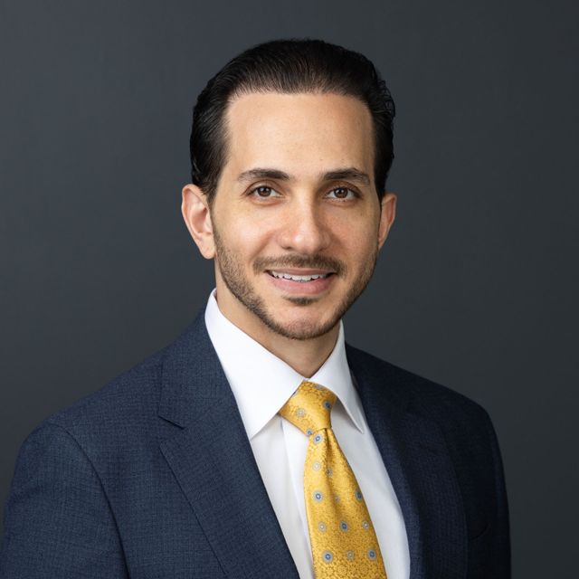 Studio headshot of a smiling professional in a navy suit, white shirt, and yellow tie against a gray background