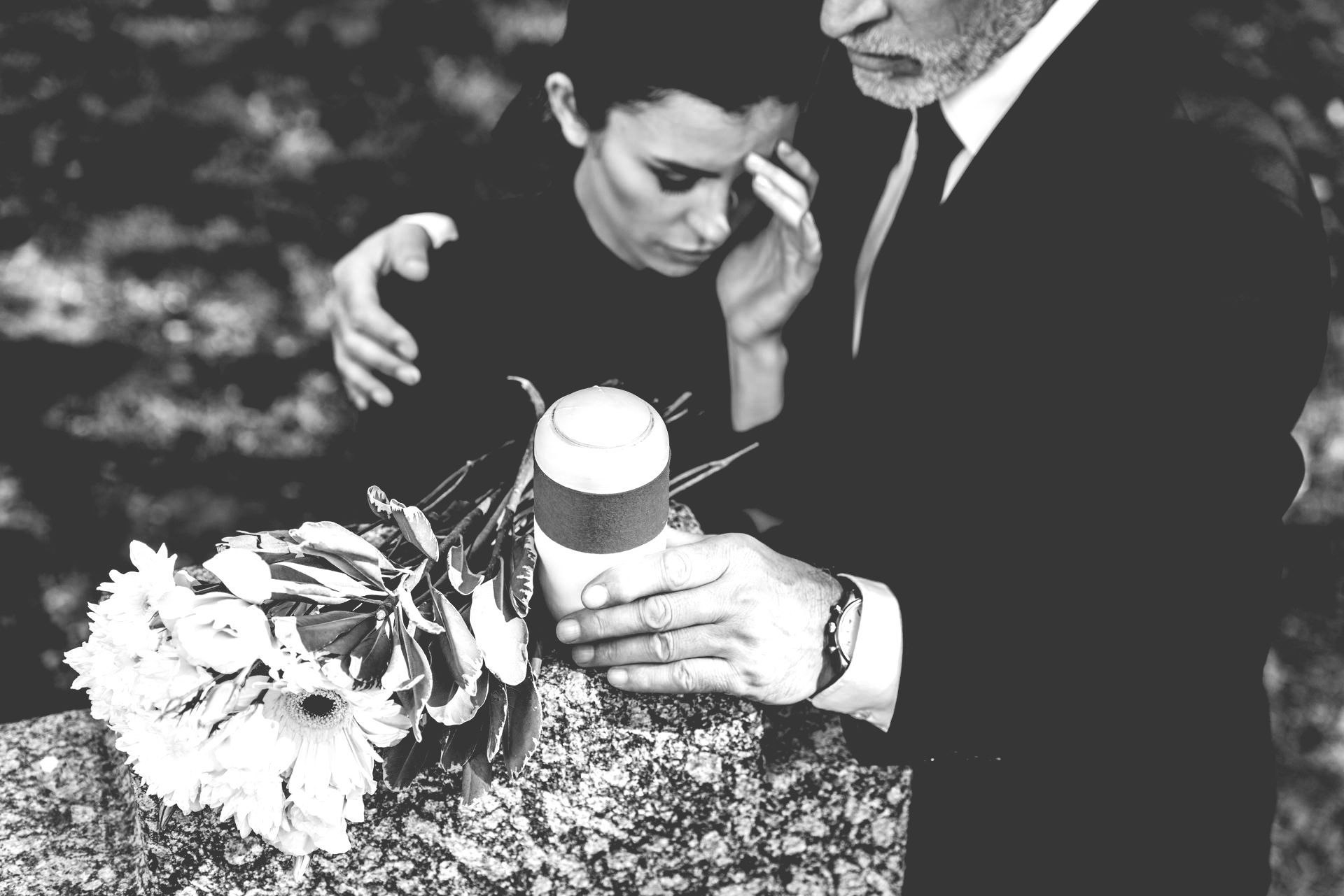 Mourner placing a hand on another’s shoulder beside a flower arrangement at a gravesite.