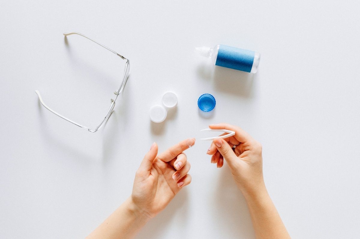 Hands holding a contact lens applicator, contact lens case, solution, and glasses on a white surface.