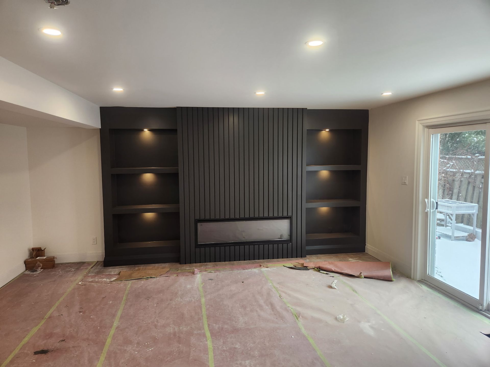 Dark gray accent wall with built-in shelves and fireplace, lit by recessed lights, in a room under construction.