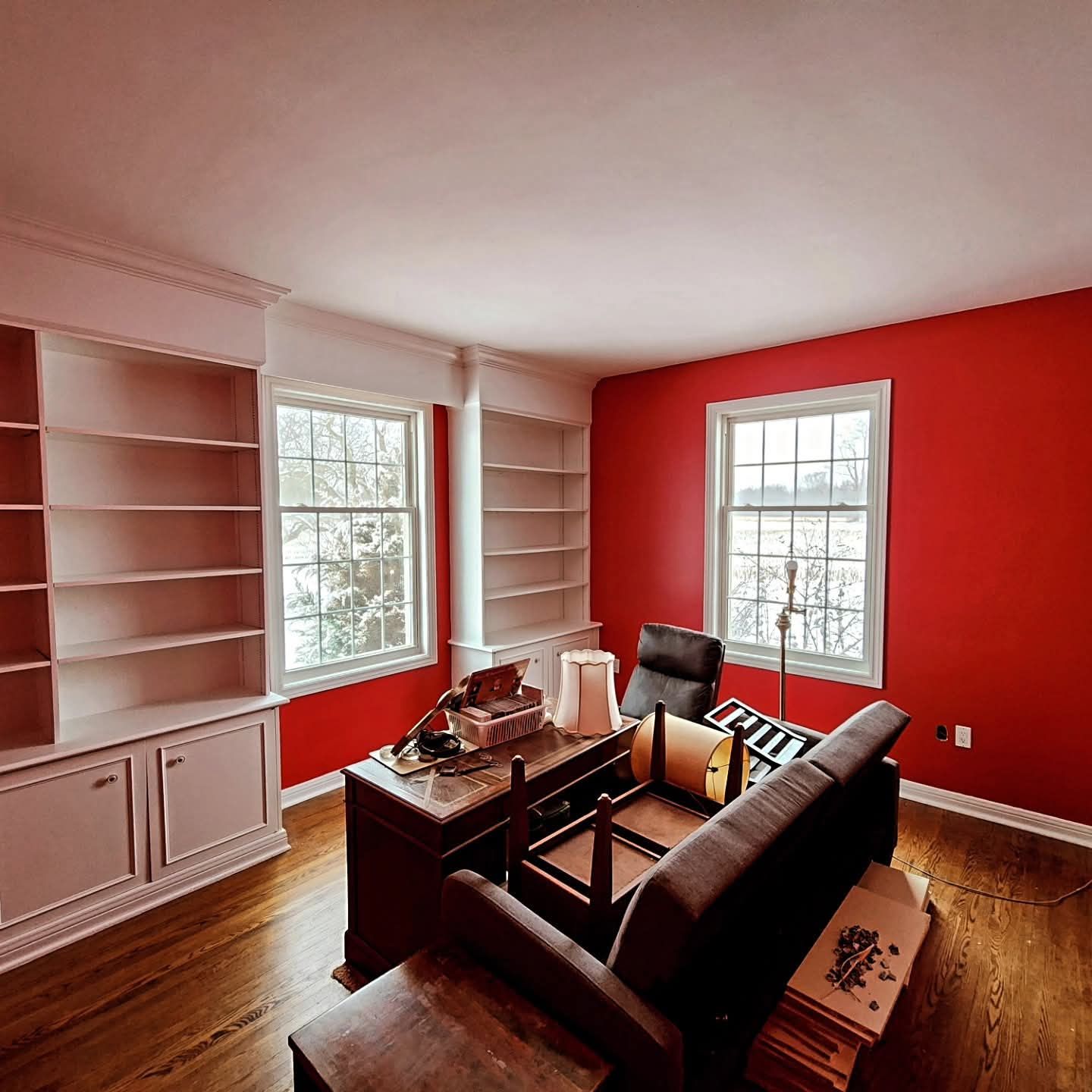 Red-walled home office with built-in bookshelves, two windows, desk, sofa, and hardwood floors.