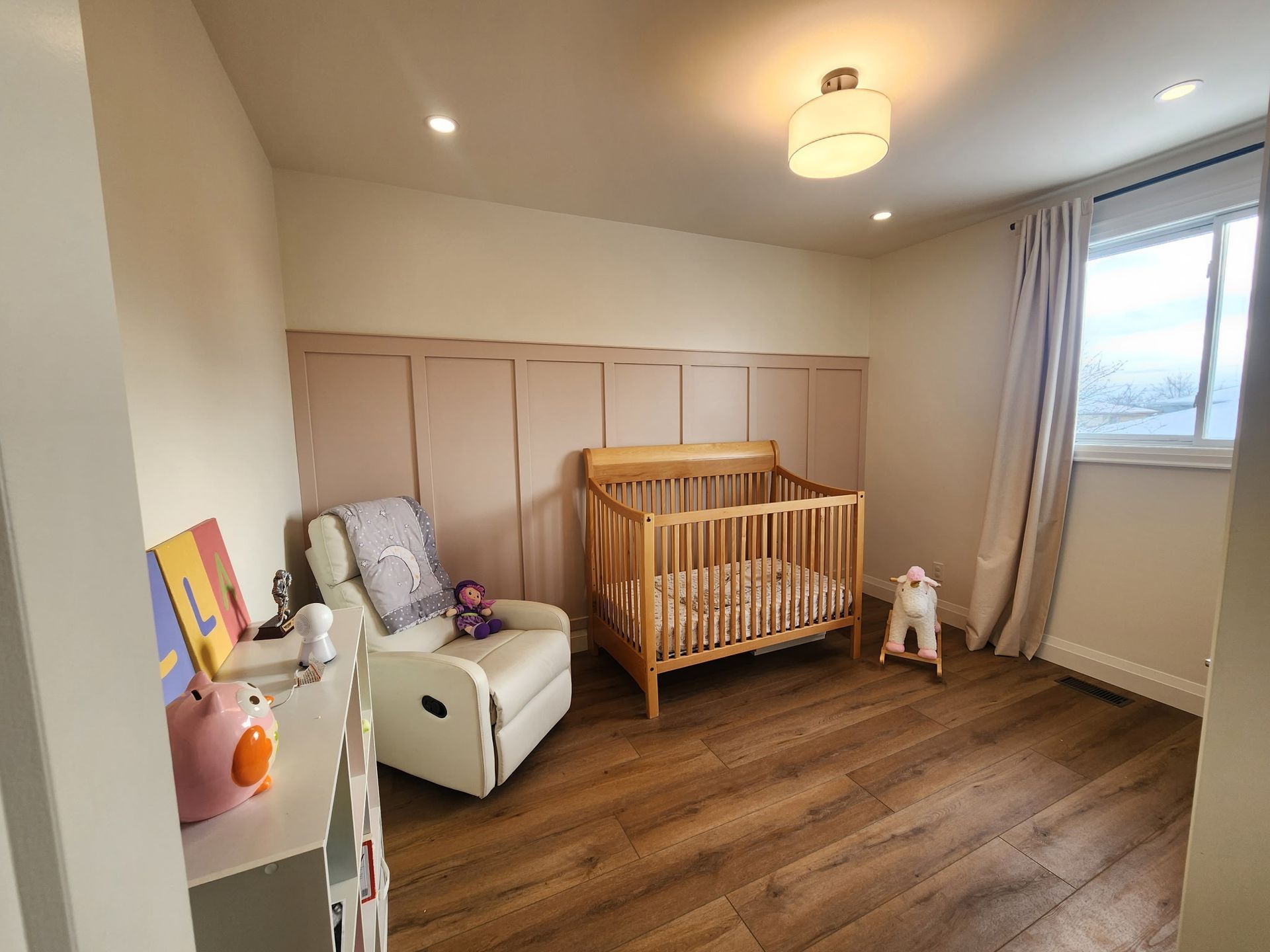 Baby room with wooden crib, rocking horse, and recliner. Light pink paneling on the wall.