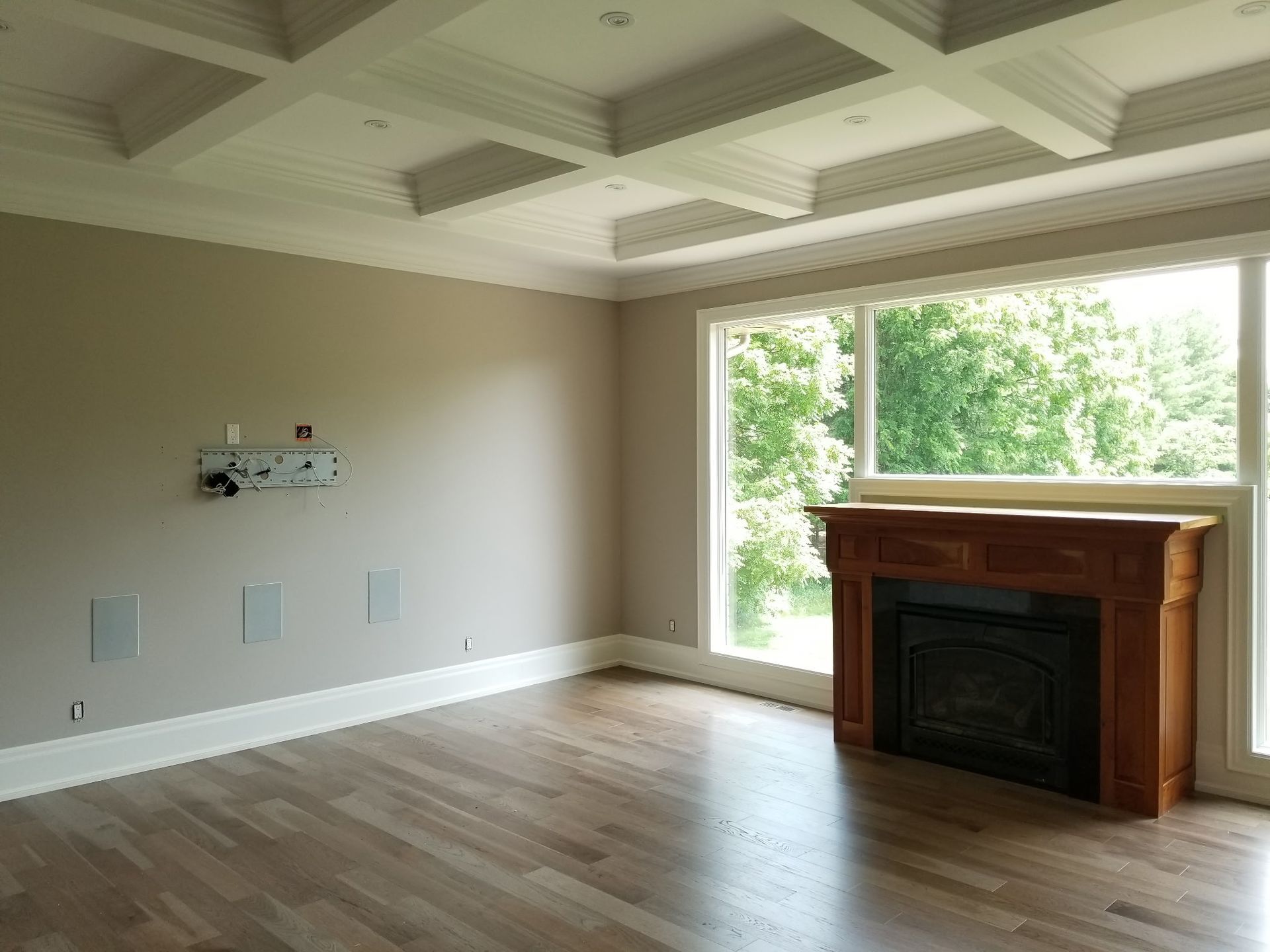 Empty room with hardwood floors, tan walls, and a coffered ceiling. A wooden fireplace is near a large window.