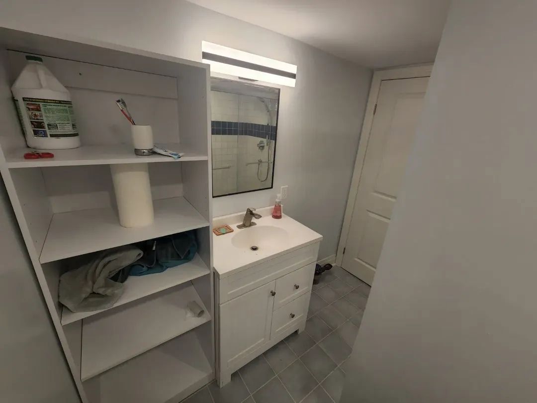 Bathroom with white vanity, open shelving, mirror, and a closed door. Light gray walls and flooring.