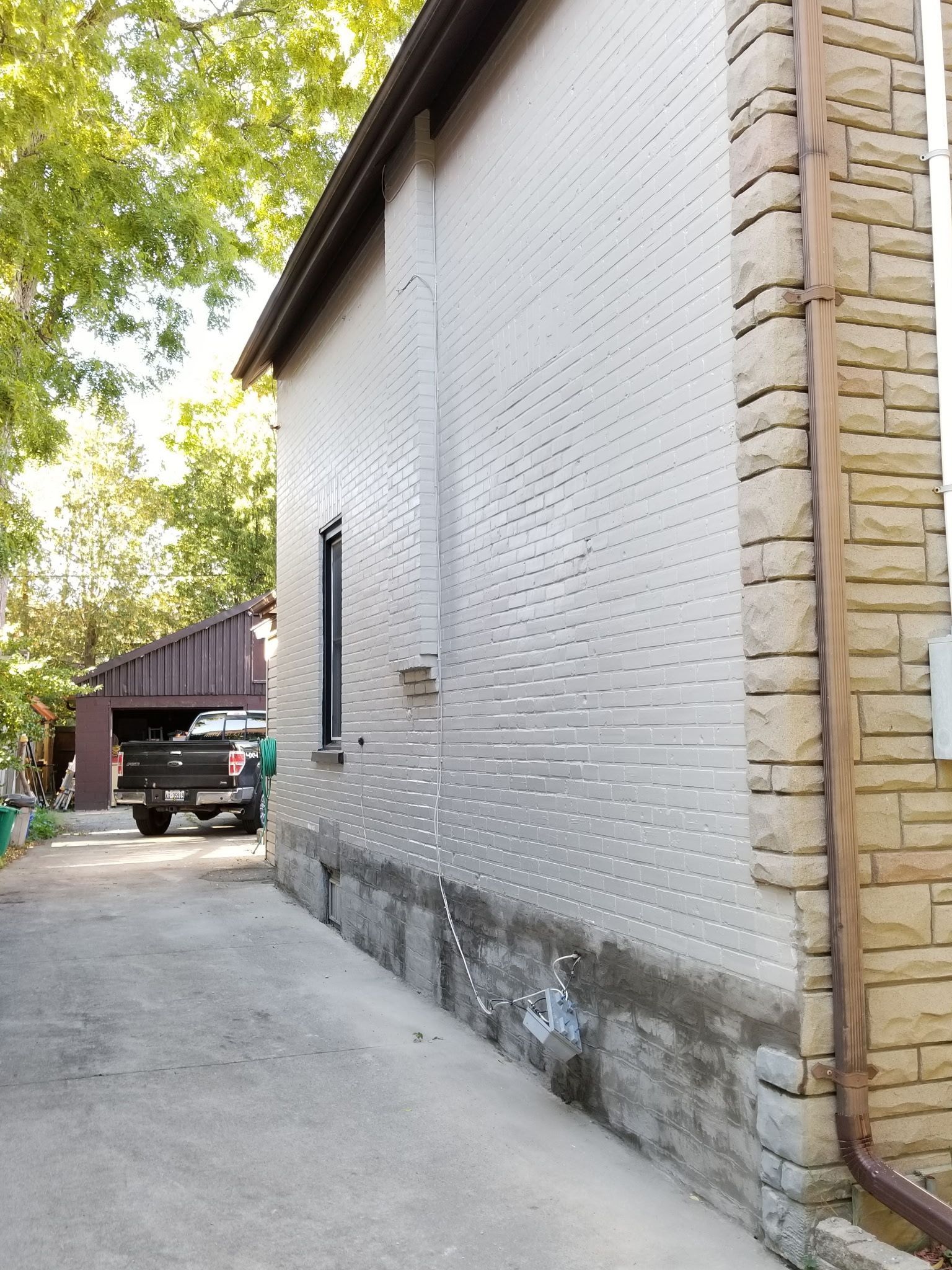 Side of a building with light gray brick, a narrow black window, and a stone facade. A driveway is in the foreground.