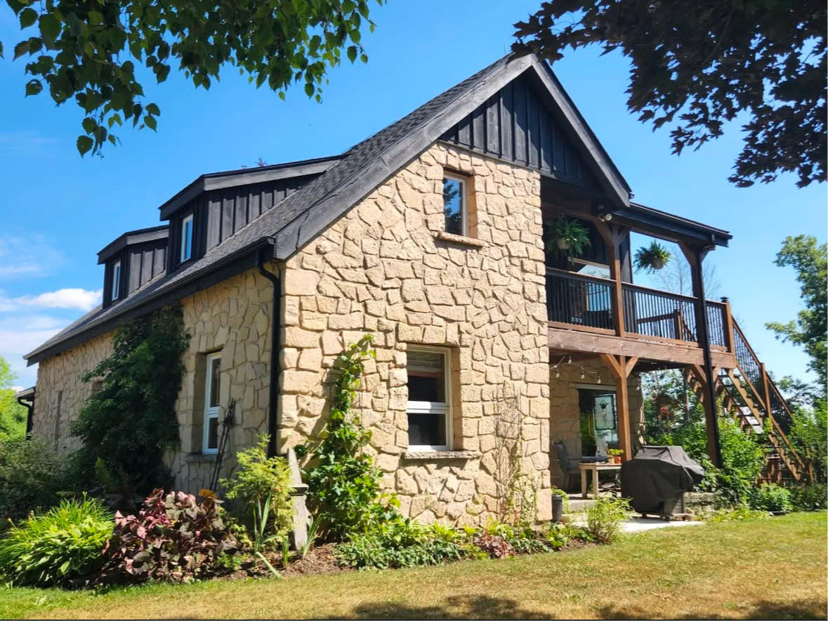 Stone house with a dark roof and a balcony. Lush greenery surrounds the house on a sunny day.