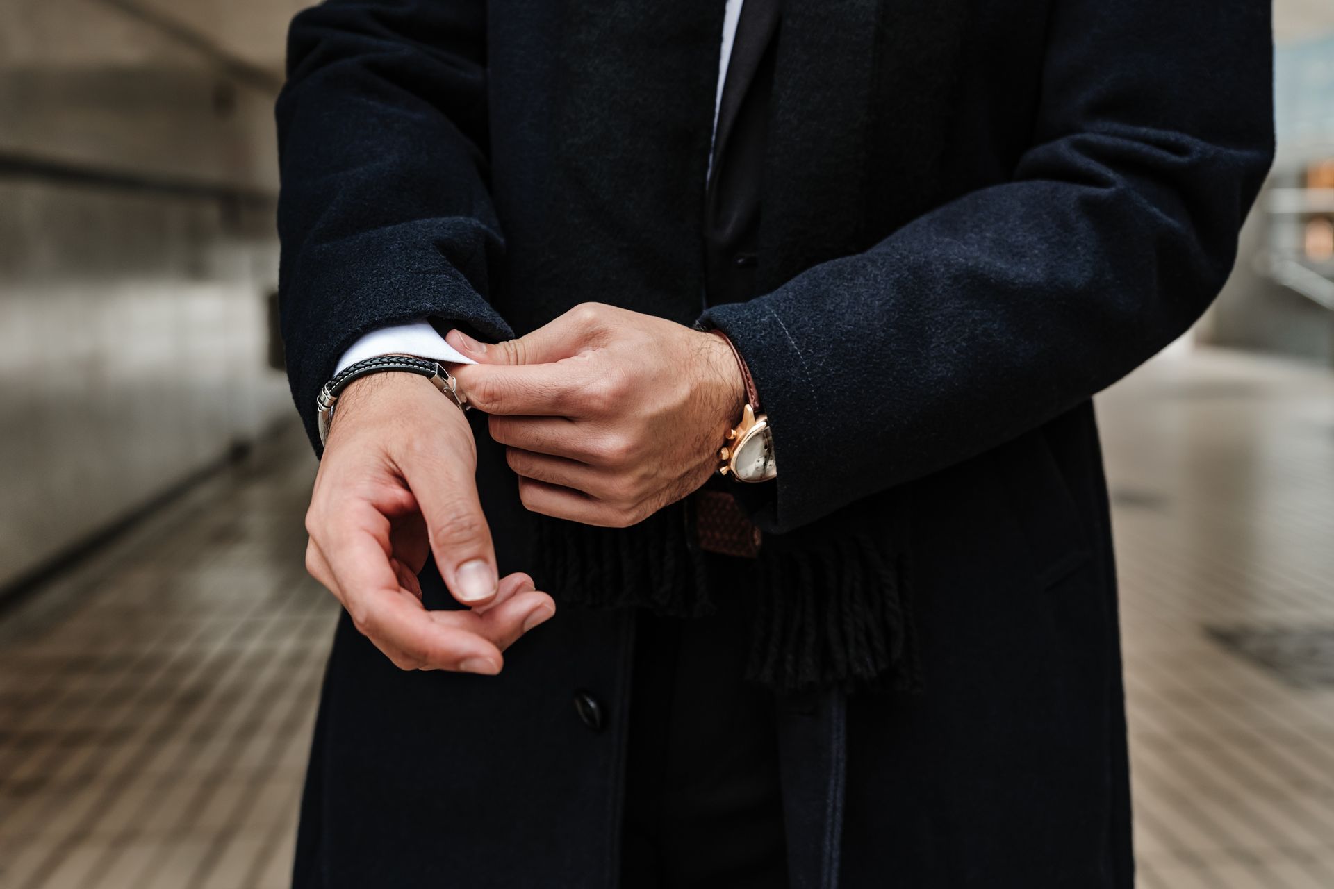 Una persona con un cappotto nero si aggiusta il polsino, mostrando un orologio. All'aperto, sfondo sfocato.