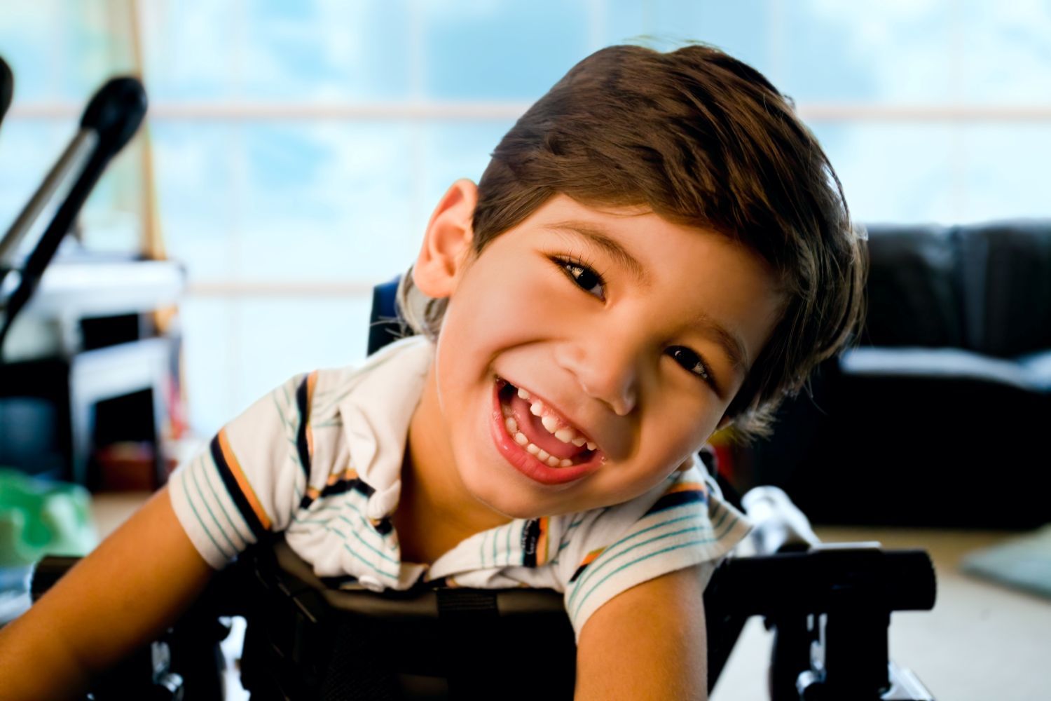 Smiling child with tilted head, wearing a striped shirt in a wheelchair indoors