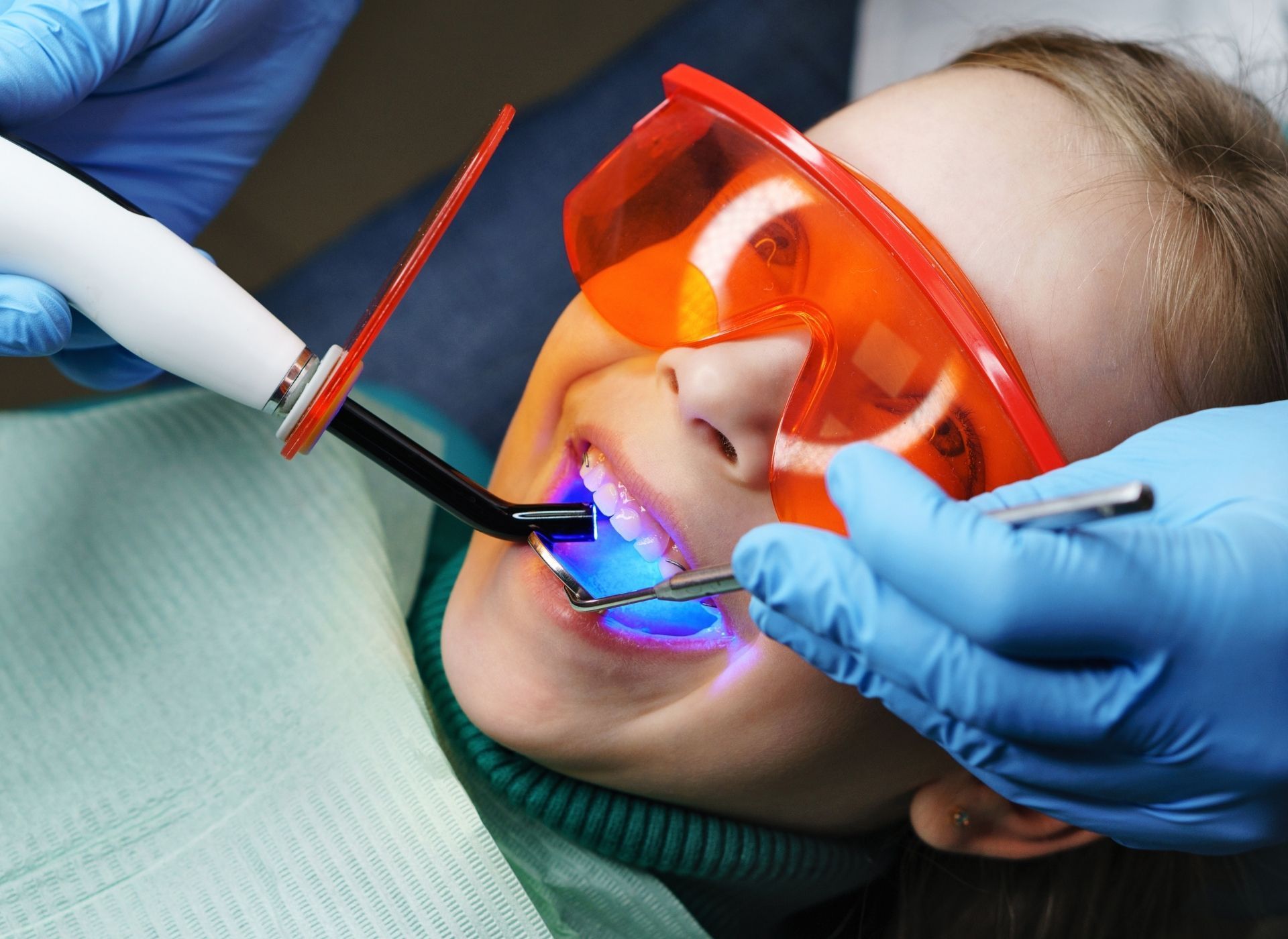 Dentist using tools to clean a child’s teeth, with orange protective glasses and blue gloves