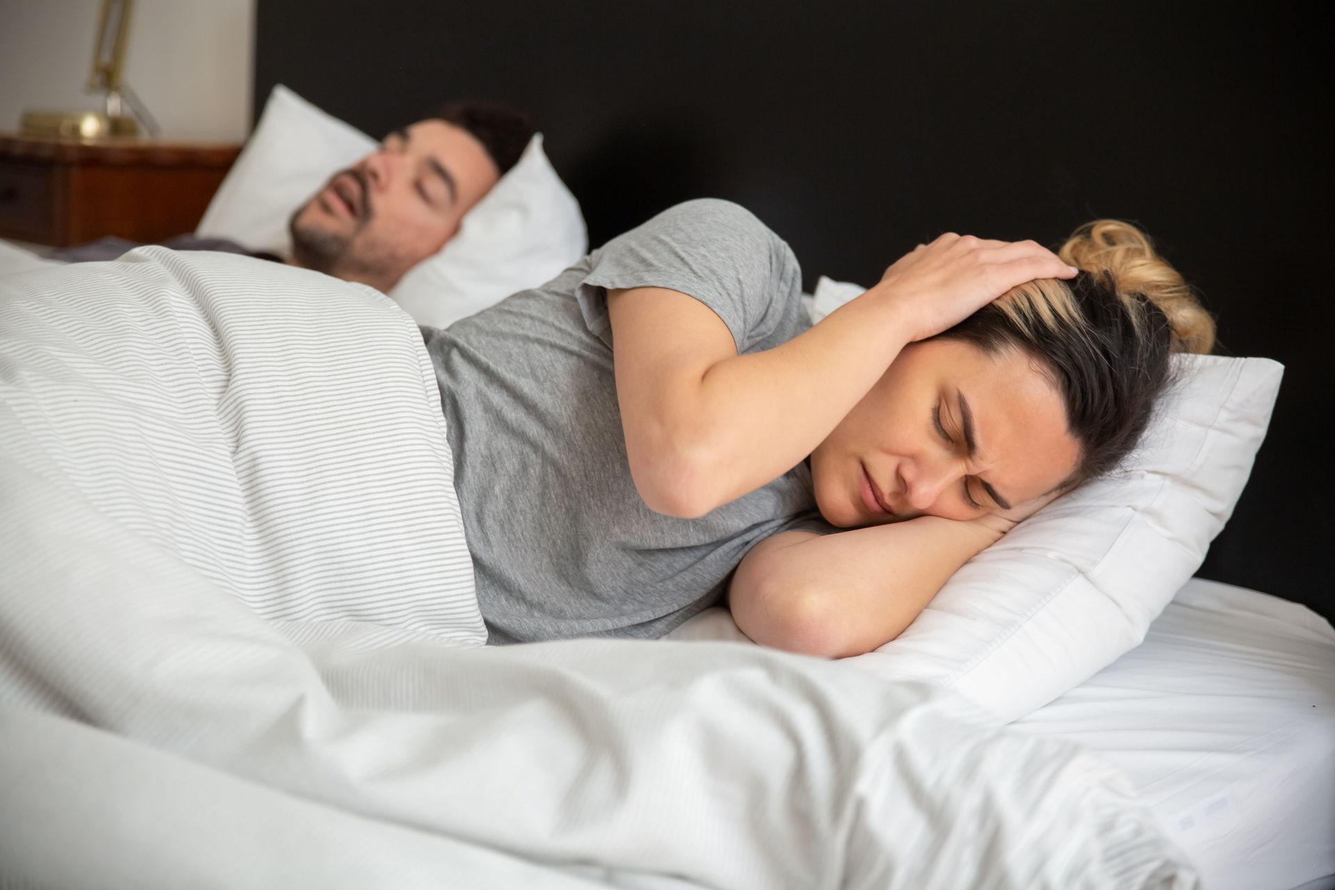 Couple sleeping in bed, man in background and woman curled under white blankets.