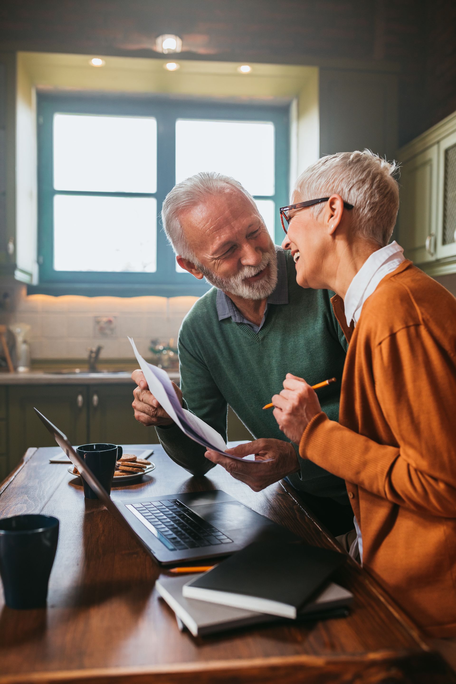 Older couple reviewing paperwork together at a table with a laptop in a bright home.