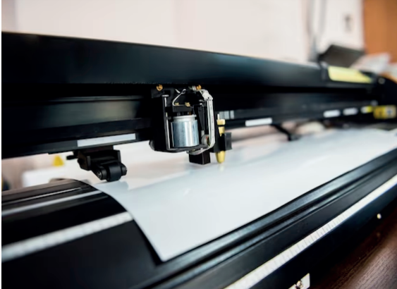 Primer plano de una cortadora de vinilo cortando una lámina blanca. La máquina es negra y automática.