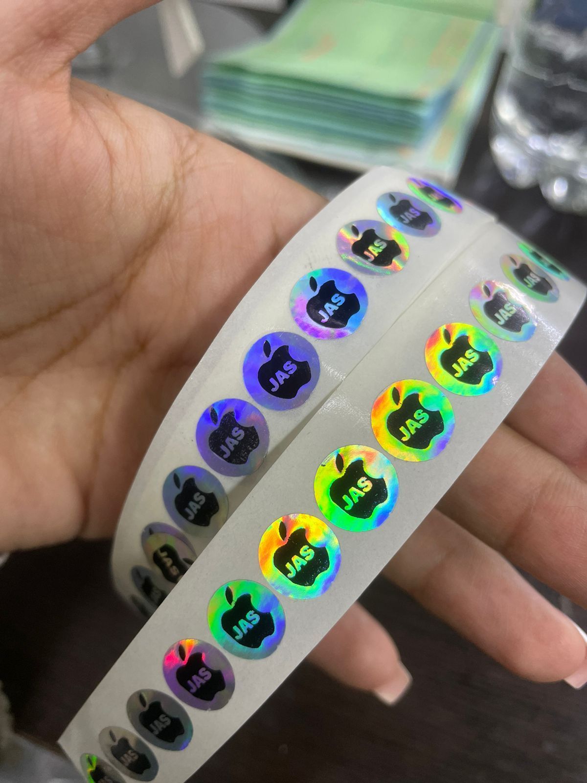 Mano sosteniendo rollos de pegatinas redondas iridiscentes con el logotipo de una manzana y el texto