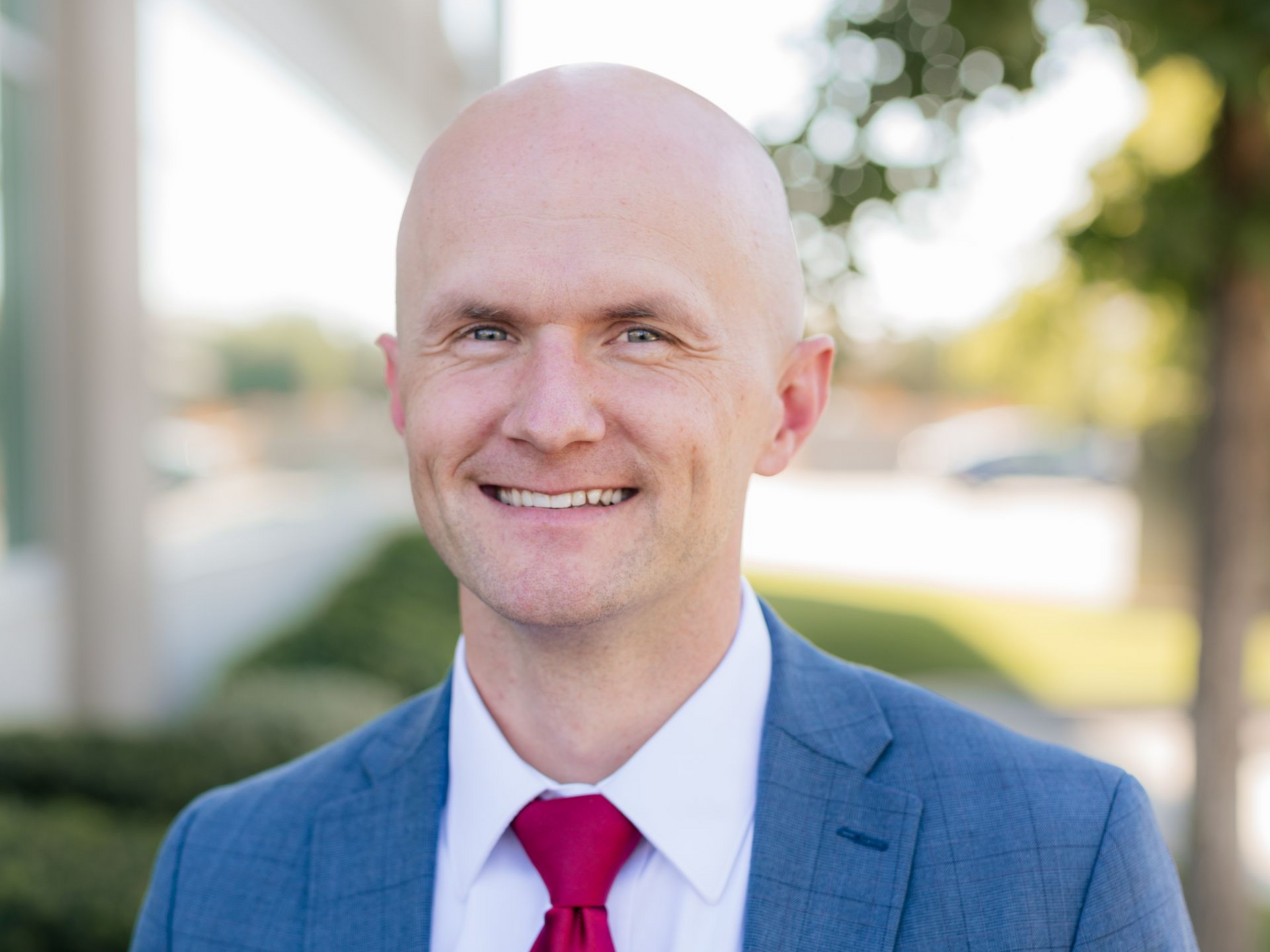Bald man in blue suit and red tie smiling, outside in front of building.