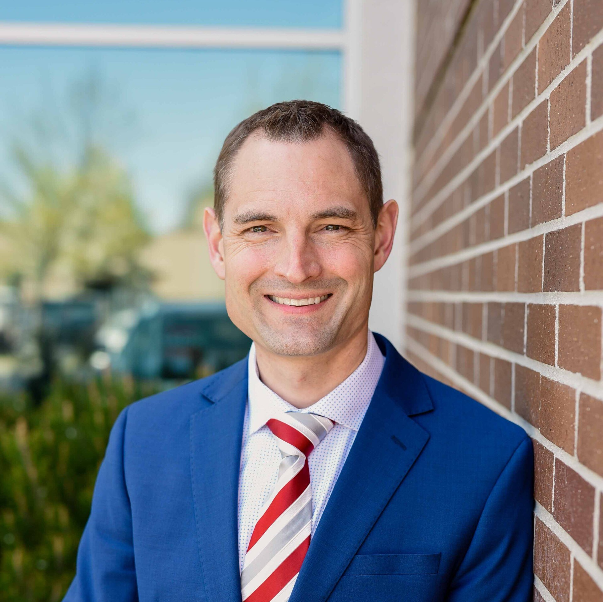 Man in blue suit smiles, leaning against a brick wall, red and gray striped tie.