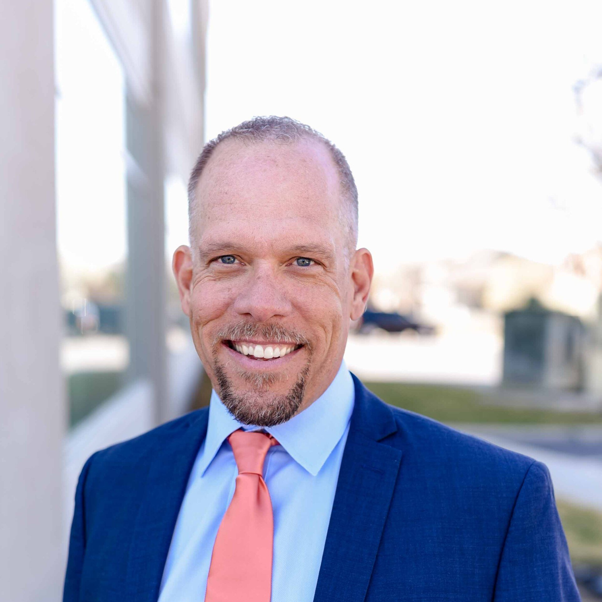 Man in blue suit and coral tie smiles outside a building.