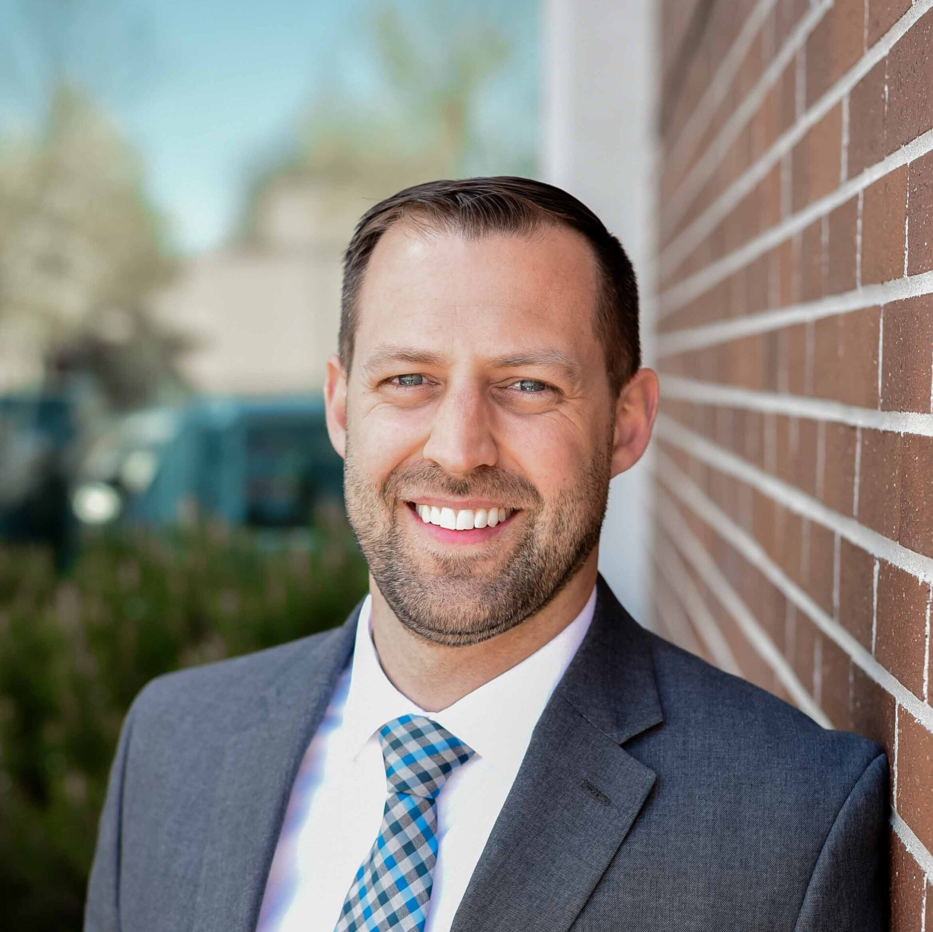 Man in a suit smiles, leaning against a brick wall outdoors.