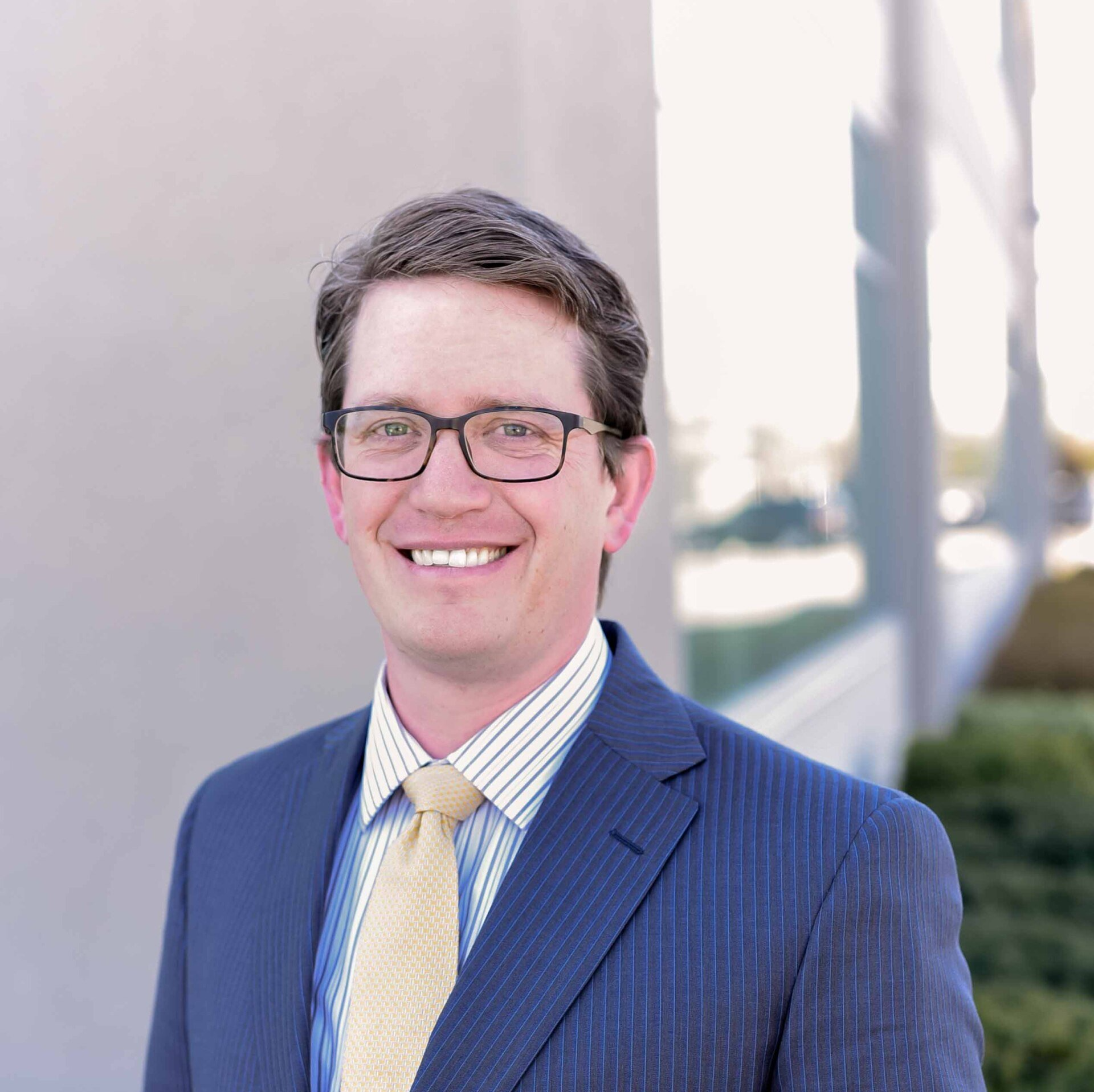 Smiling man in a blue pinstripe suit and glasses, standing outside a building.