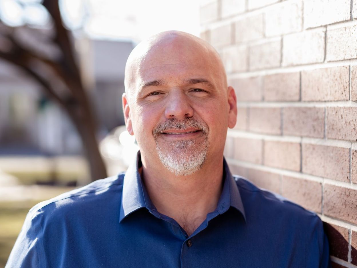 Bald man smiles at the camera while leaning against a brick wall; he has a goatee and wears a blue shirt.