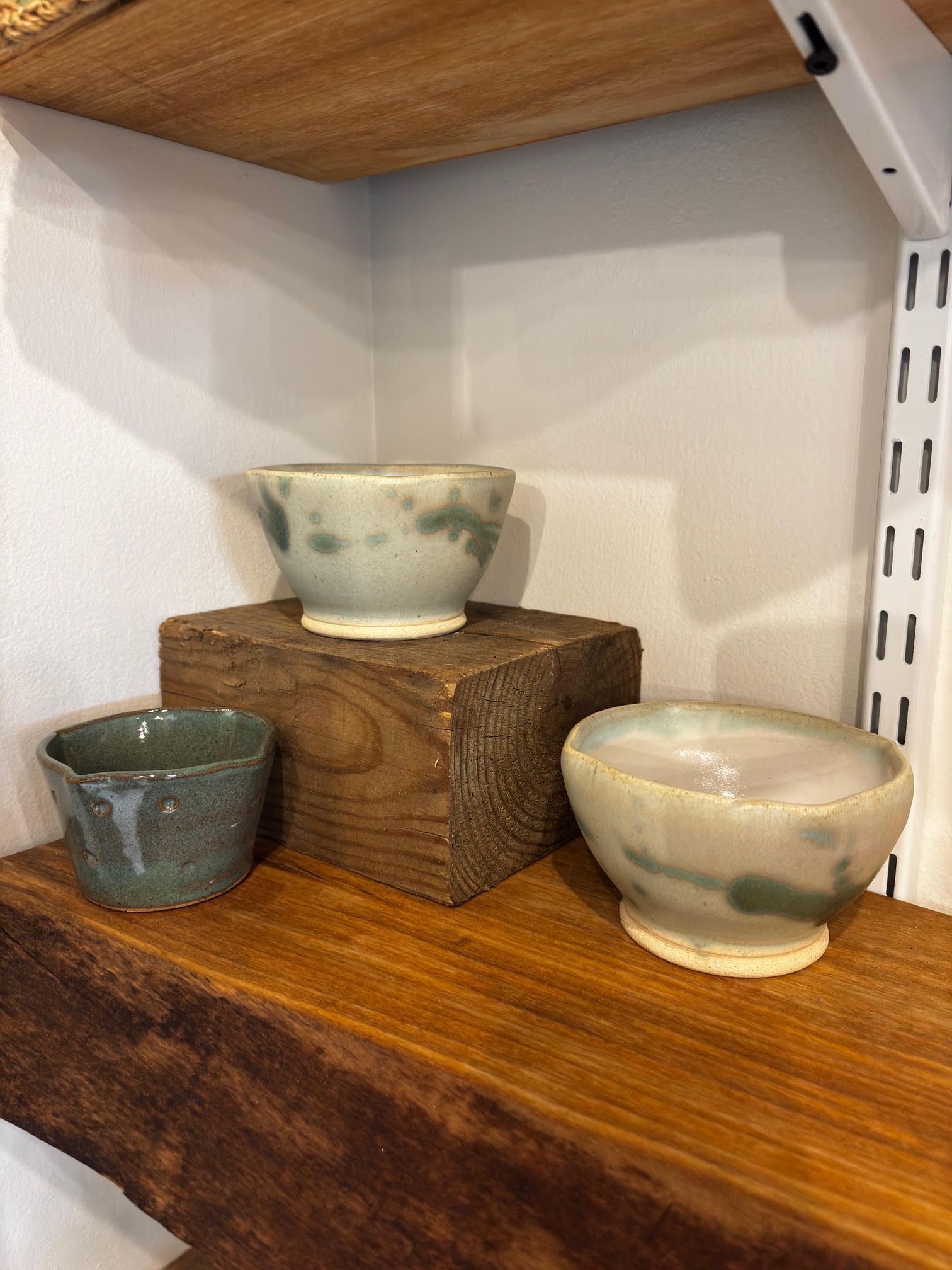 Three small ceramic bowls on stacked wooden blocks on a shelf, in muted green and cream tones