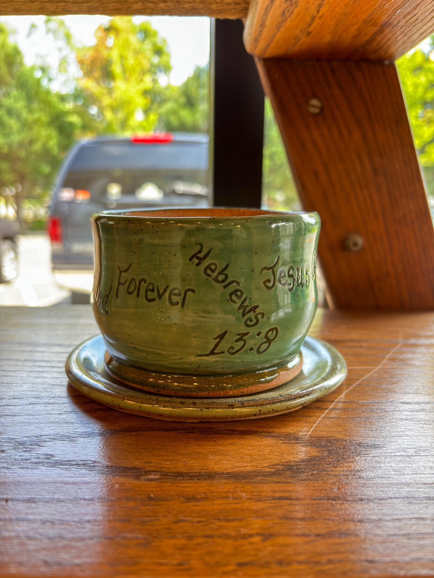 Hand-painted green ceramic cup with text, on a wooden table near a blurred car outside