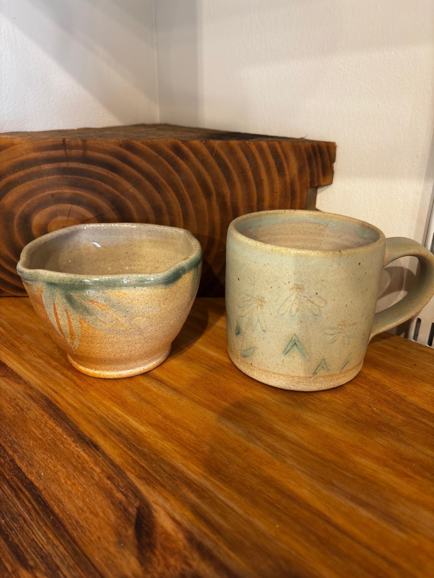 Two handmade ceramic mugs on a wooden shelf, one speckled beige and one blue-green.