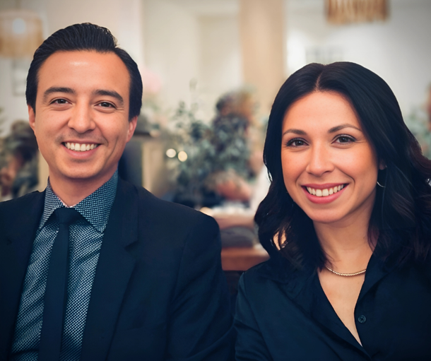 Two smiling people in dark business attire seated indoors at a formal event