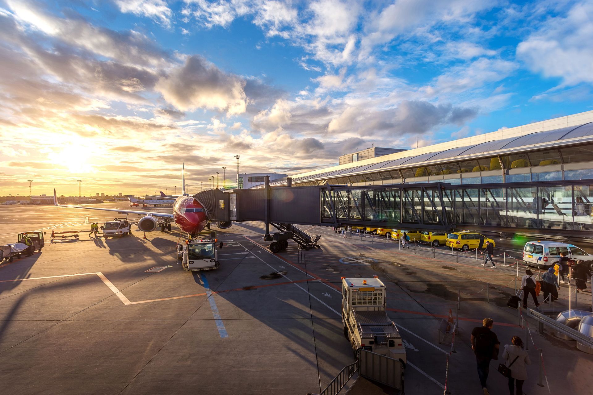 Airport terminal exterior at sunset with planes, ground vehicles, and travelers on the tarmac