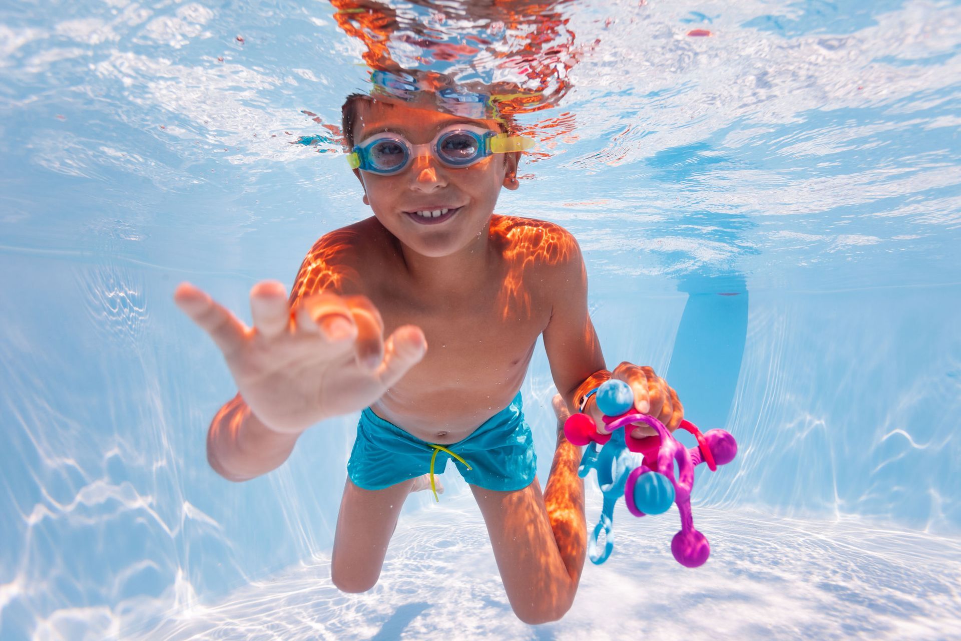 Boy underwater, wearing goggles and swim trunks, reaching out and smiling, holding a toy.