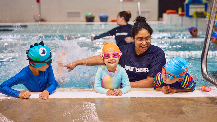 A swim instructor helps two toddlers in a pool, while their mothers watch.