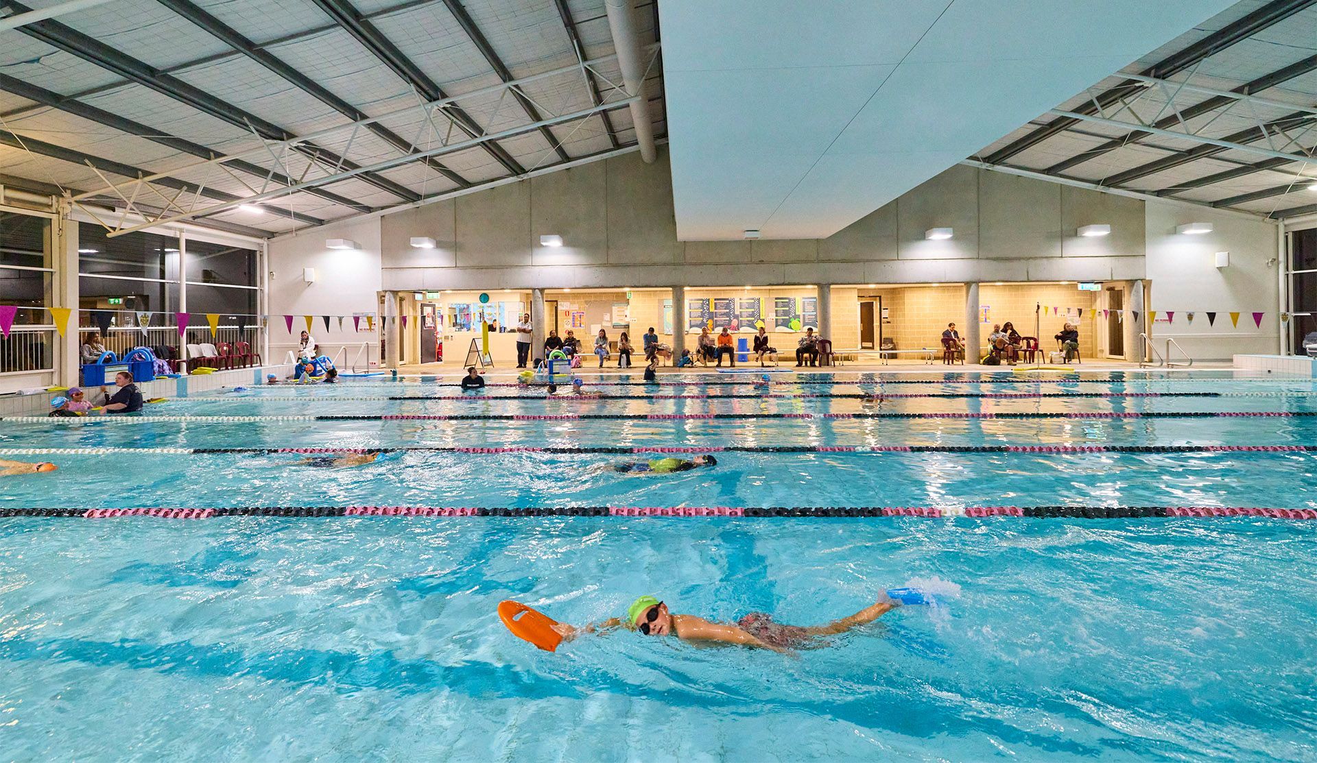 A large indoor swimming pool with lanes running horizontally and a parent seating area at the back