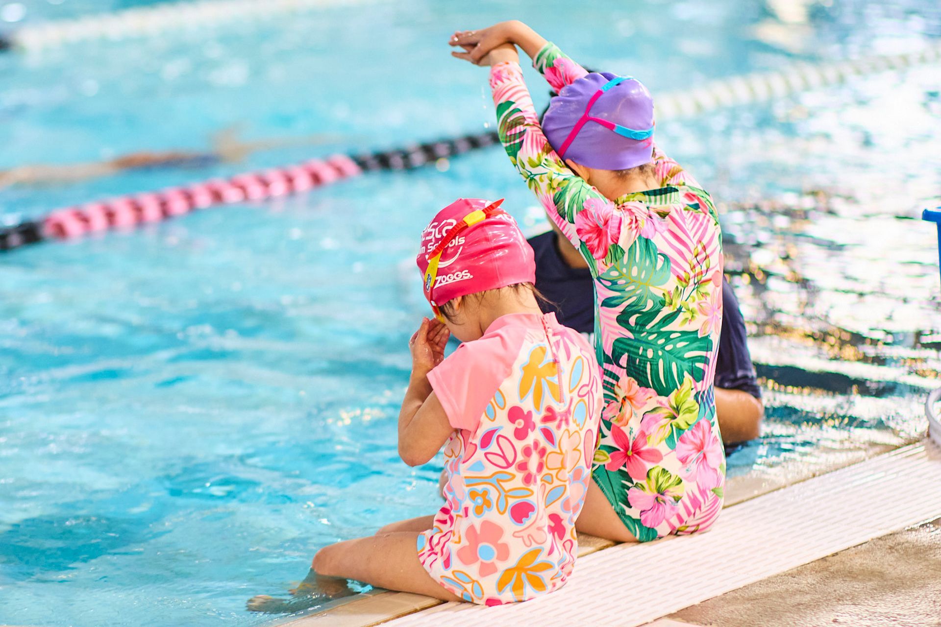 Two girls in swimsuits and caps sit by a pool, one raising arms, floral patterns, pink and blue.