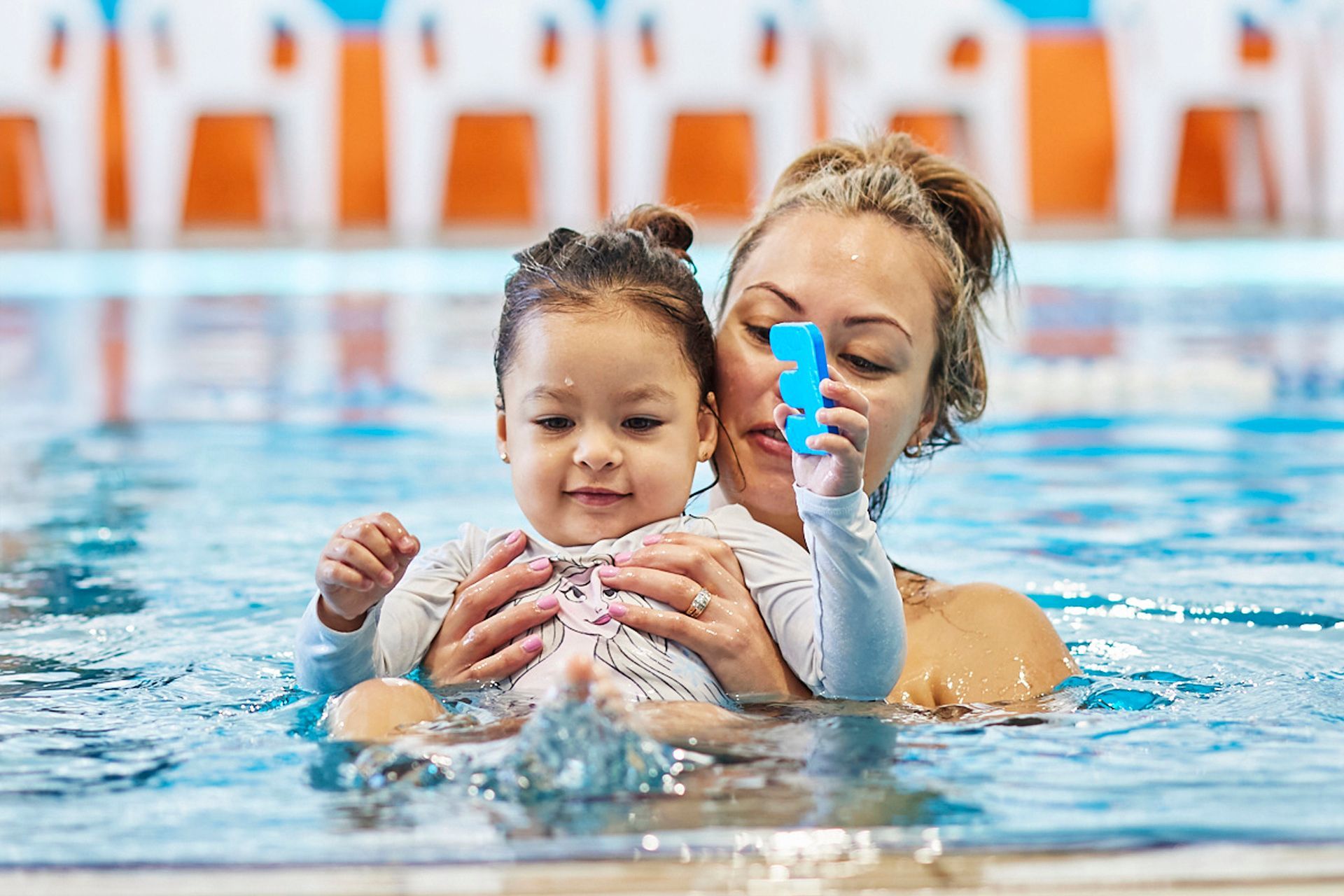 Woman and toddler in a pool, smiling. Woman holds a blue toy; white pool setting.