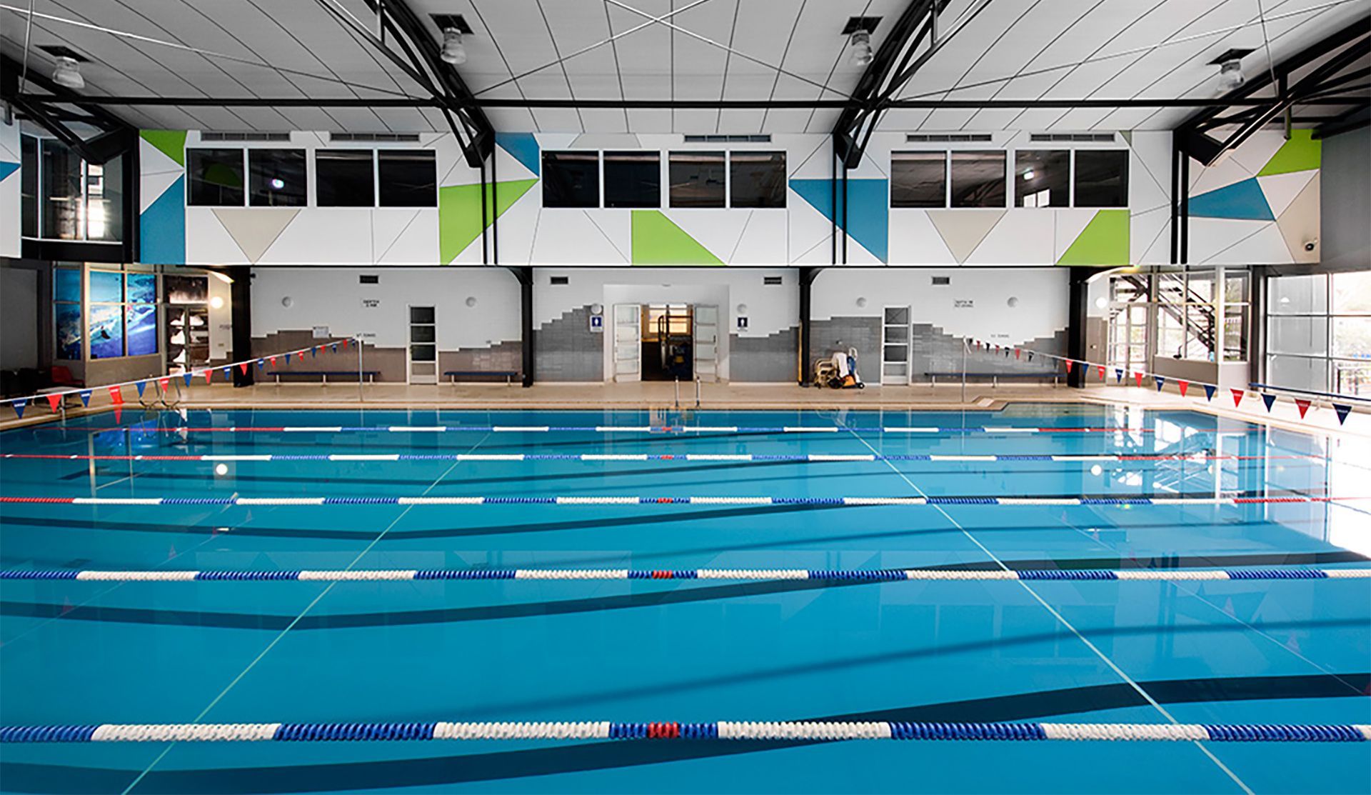 Indoor swimming pool with blue water, lane markers, and a geometric design on the wall.