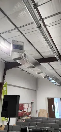 Industrial ceiling with exposed ducts and vents above rows of gray chairs in a hall