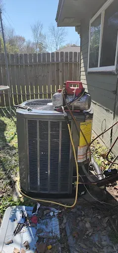 Outdoor HVAC unit with hoses and a red gauge on top beside a house and wooden fence.