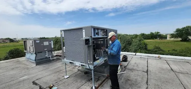 Worker inspecting rooftop HVAC unit beside another unit on a flat roof under a cloudy sky