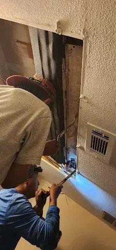Technicians working inside a wall cavity, using a flashlight and tools near exposed wires in a cramped utility area