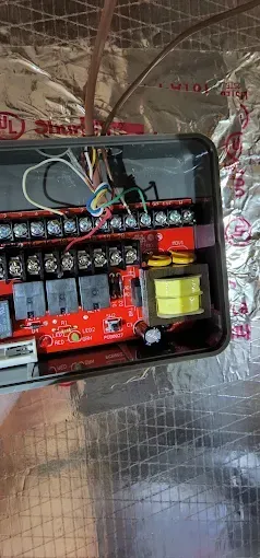 Open electrical control box with wires, relays, and switches mounted on a wall.