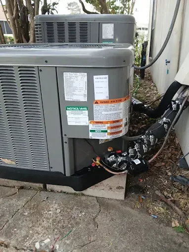 Two gray outdoor HVAC units beside a house, with hoses and labels on the larger unit.