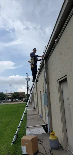 Worker on a ladder cleaning the side of a building, with tools and boxes below.