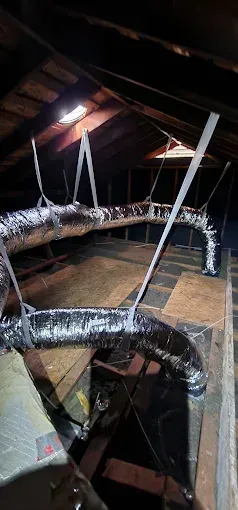 Insulated HVAC ducts and metal supports in a dark attic with exposed rafters.