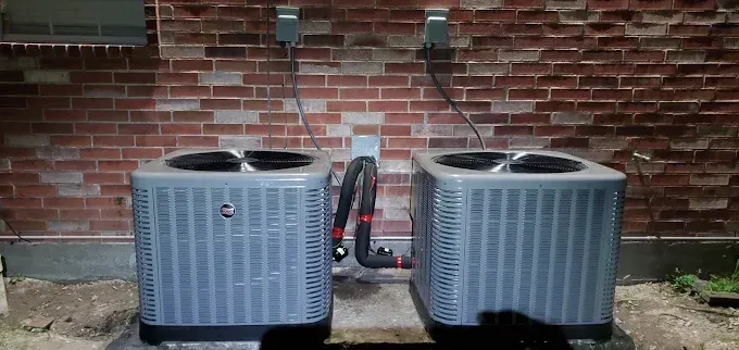 Two gray outdoor HVAC units beside a brick wall, with connecting pipes between them.