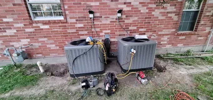 Two gray outdoor AC units beside a brick wall with maintenance tools on the ground