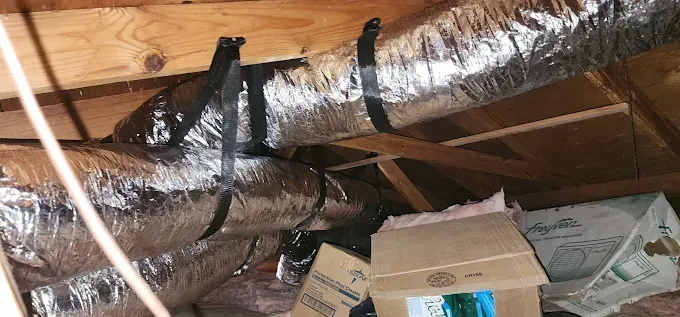 Insulated silver ductwork with black tape in an unfinished attic above boxes and ceiling beams